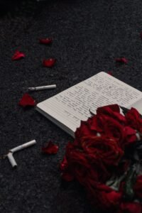 Home A moody photo featuring red roses, scattered cigarettes, and an open notebook on a dark background.